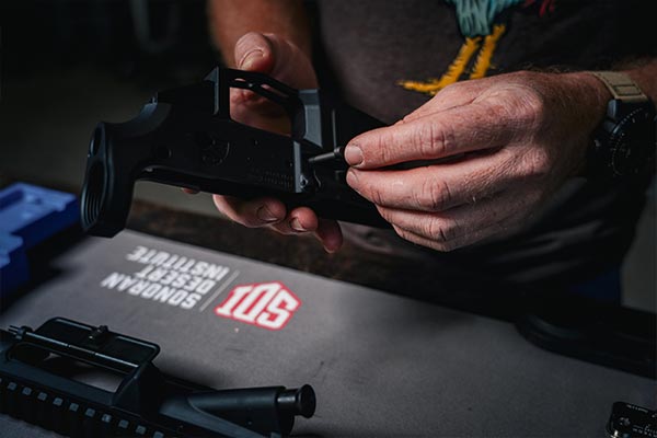 Close-up of hands working on an AR-15 lower receiver during SDI gunsmithing training on a branded bench mat.