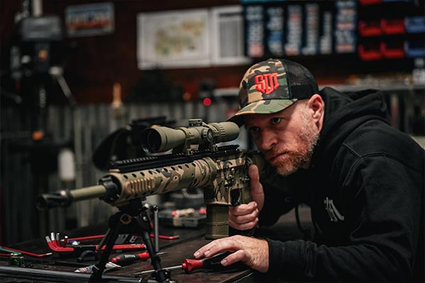 Man in SDI cap aiming a custom AR-style rifle with scope on a workbench in a gunsmithing shop.